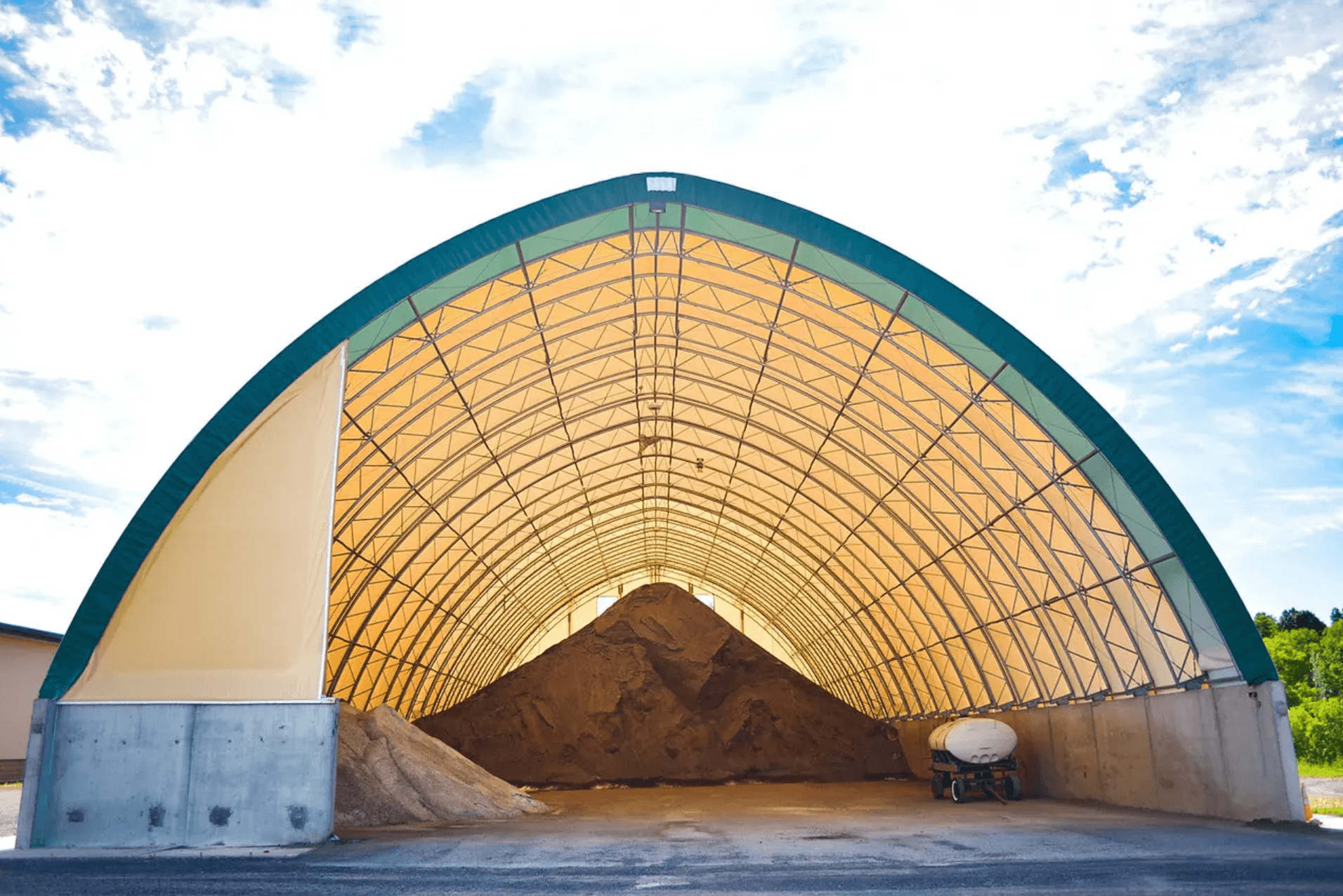 Large arch-shaped storage building with a translucent roof, housing a massive pile of brown granular material, with a white tanker truck partially visible on the right.
