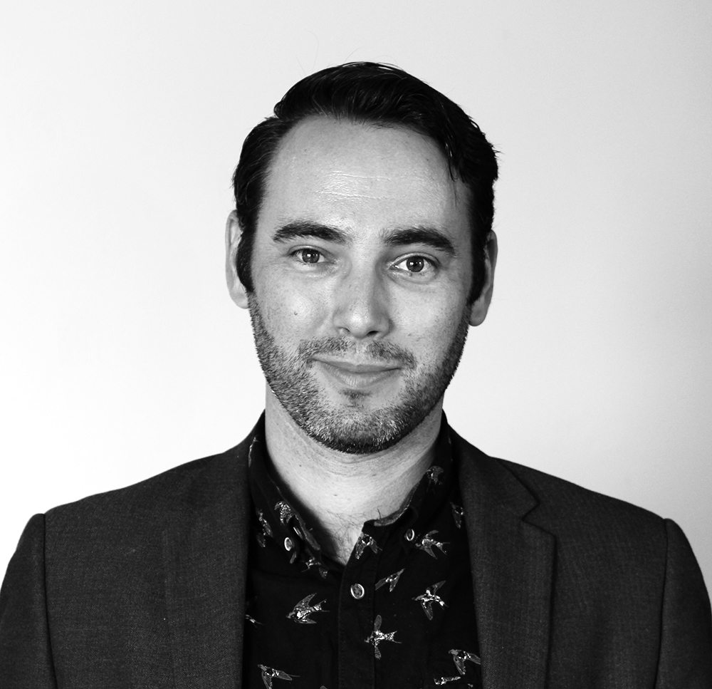 Black and white portrait of a man with short hair and a light beard, wearing a jacket over a shirt with bird patterns.