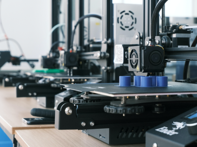 A 3D printer with a black frame printing a blue object on a heated print bed, with other 3D printers and filament spools in the background.