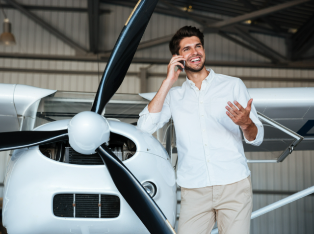 Smiling man talking on a mobile phone while gesturing with his hand, standing in front of a small propeller aircraft inside a hangar.