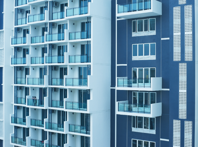 Modern apartment building with a blue and white facade, featuring balconies on each floor and a person standing on one of the balconies.