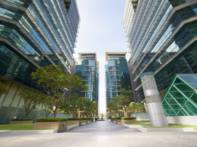 Symmetrical view of a pedestrian walkway between contemporary glass high-rise buildings with trees and benches under a clear blue sky.