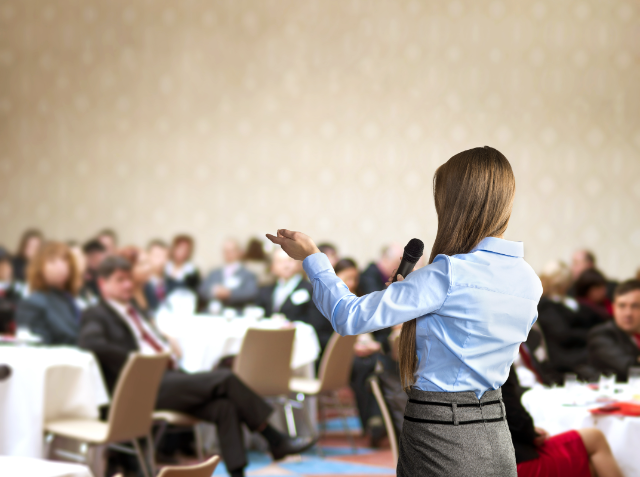 A woman presenting at a conference with a microphone, gesturing to an audience seated at tables.