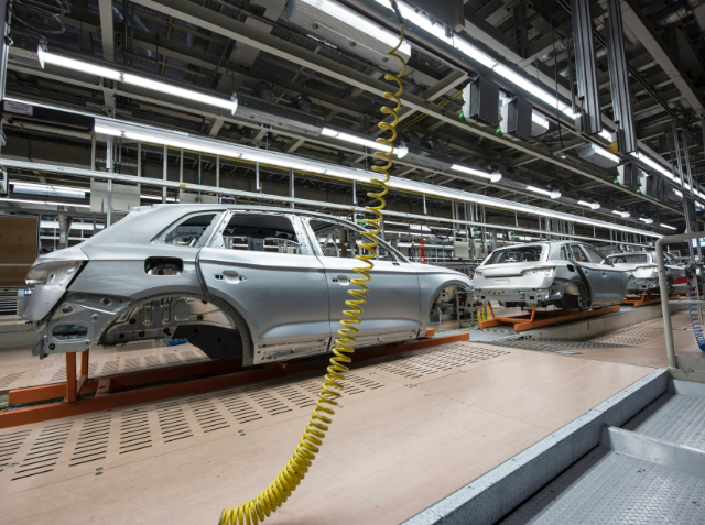 Partially assembled car bodies on an assembly line in a vehicle manufacturing plant, with a yellow coiled cable in the foreground and various lines and cables overhead.