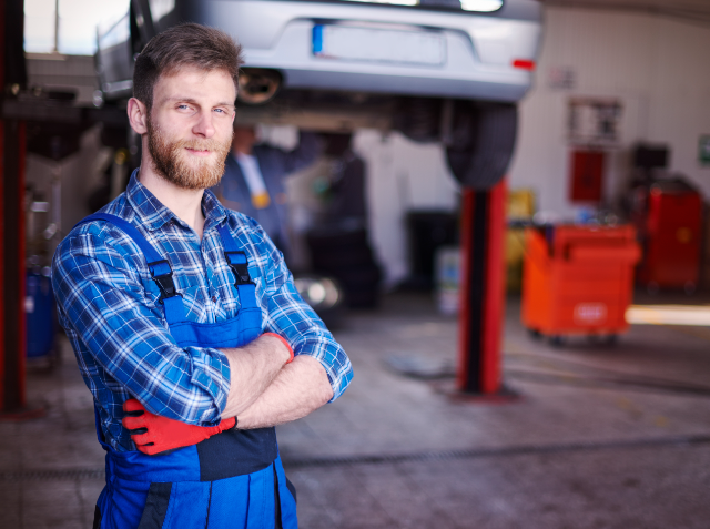 Confident mechanic with a beard standing with arms crossed in an auto repair shop, with a car on a lift and tools in the background.