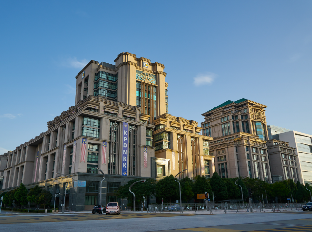 A large, multi-story building with a central clock tower and decorative elements, situated on a city street under a clear blue sky.