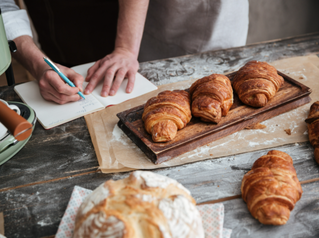 A person in an apron writing on a notepad next to a wooden board with five freshly baked croissants, in a kitchen setting with baking equipment.