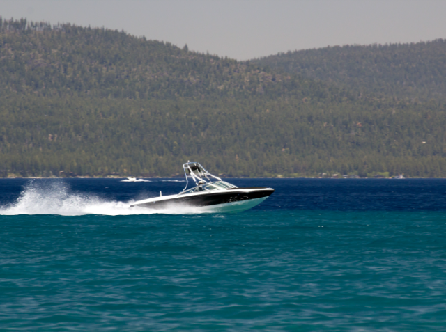 A speedboat cruising on a blue lake with a wake behind it, with forested mountains in the background under a clear blue sky.
