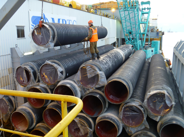 A worker in safety gear stands beside a stack of large black corrugated pipes on an industrial ship deck.