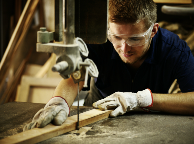 A focused man with safety goggles and gloves using a band saw to cut wood in a workshop.