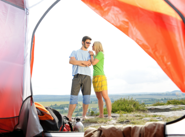 A couple embracing outside a camping tent with camping gear in the foreground and a scenic landscape in the background.