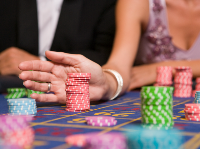 Close-up of a stack of red poker chips on a casino table with players' hands visible and other colorful chips in the background, indicating an ongoing game.