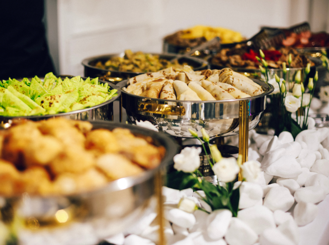 A buffet table with an assortment of dishes in metal serving trays, including baked goods and green lettuce, decorated with white flowers, at a festive event.