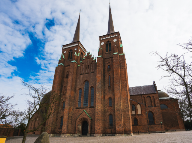 A large brick Gothic church with twin spires and pointed arch windows under a partly cloudy sky, with a bare tree and paved ground in the foreground.