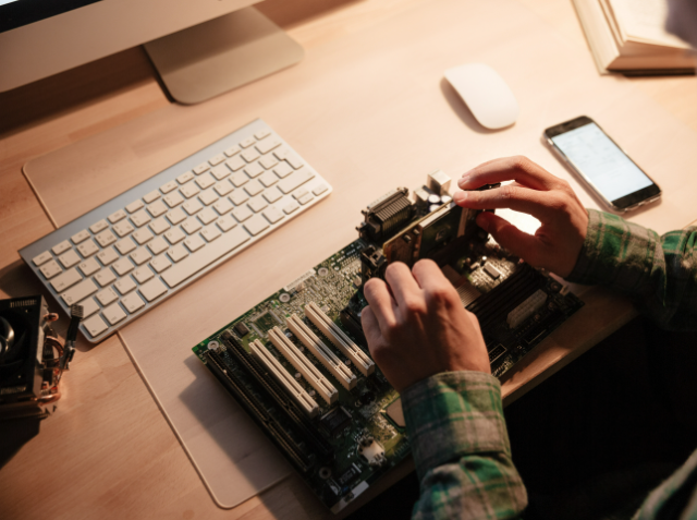 Person working on a computer motherboard, holding a component above it, with a keyboard, mouse, smartphone, and camera lens on the desk.