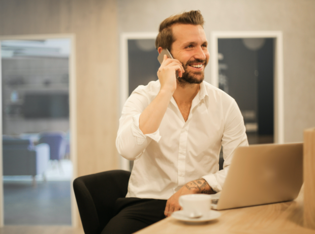 Smiling man in a white shirt talking on a mobile phone while sitting at a table with a laptop and a coffee cup, with a blurred office background.