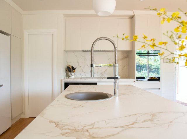 Modern kitchen with white cabinetry, marble countertops with veined pattern, undermount sink with high-arc faucet, window with greenery view, and yellow flowers in the foreground.