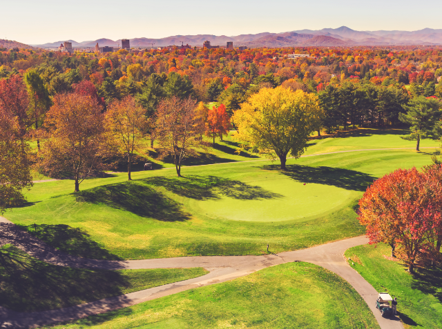 Autumnal view of a golf course with colorful trees, a winding cart path, distant buildings, and a mountain range under a clear blue sky.