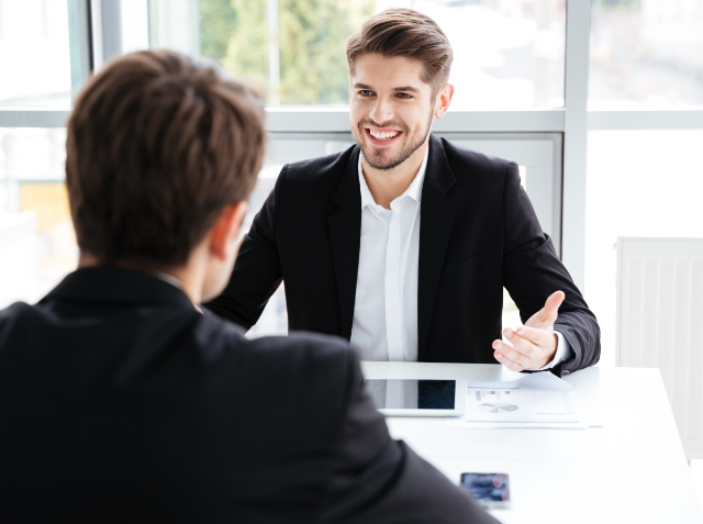 A man in a business suit smiling and gesturing during a conversation with another person whose back is to the camera.