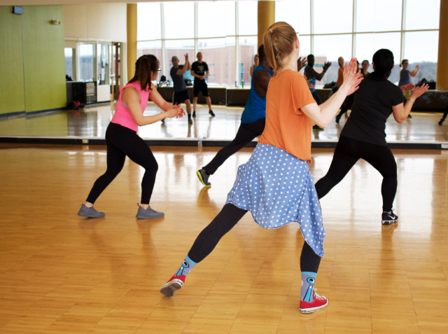 A young girl in a blue polka dot skirt and orange shirt participates in a dance class with adults in a room with wooden floors and mirrored walls.