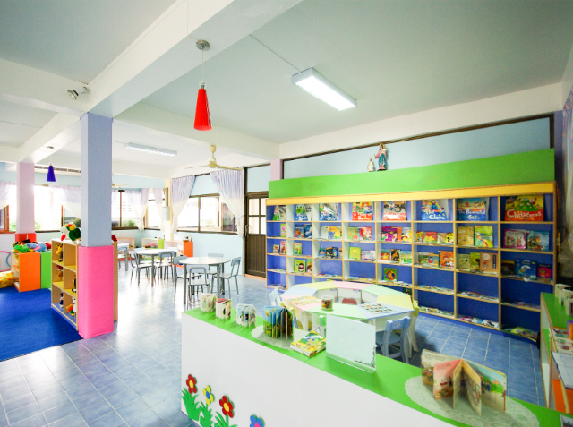 Colorful classroom interior with shelves of board games, small tables with chairs, and brightly colored walls and hanging lamps.