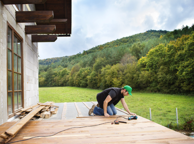 A person is working on a wooden deck using a power tool, with construction materials around them, next to a building with a forested hill in the background.