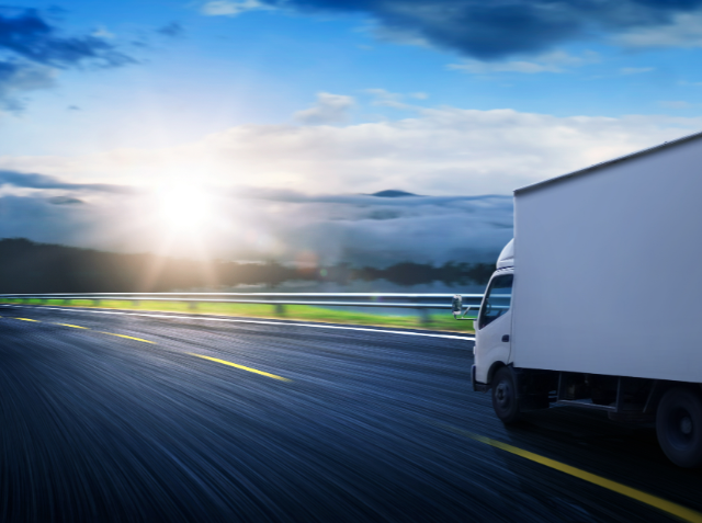 A white delivery truck in motion on a highway with a motion blur effect, with the sun shining through clouds over a hilly landscape at sunrise or sunset.