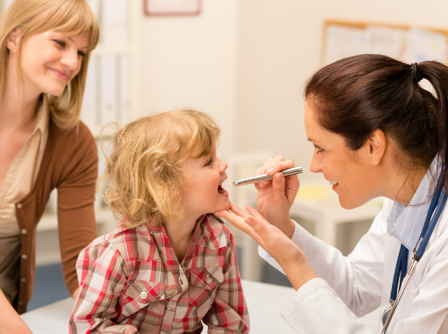 A pediatrician examines a young child's throat with a penlight as a smiling woman, likely the mother, looks on in a medical office.