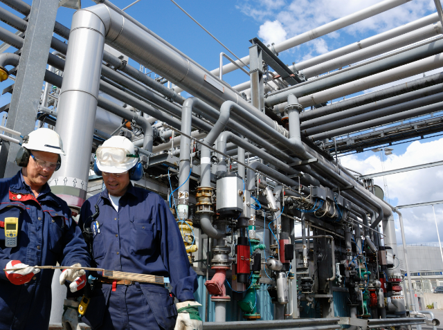 Two industrial workers in hard hats and safety gear inspecting equipment at a complex industrial plant with numerous pipes and machinery.