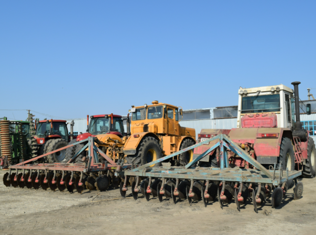 Three tractors with farming implements parked in a row on a dirt surface, with additional agricultural equipment in the background.