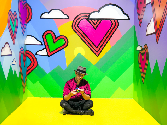 Person in a hat and shiny pink jacket sitting cross-legged on the floor in a colorful room with a cartoon-like heart and cloud mural.