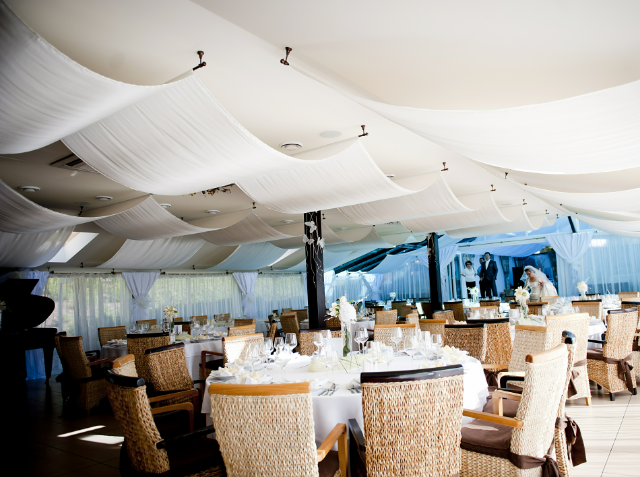 An elegant indoor event space with round tables set for dining, wicker chairs, and white draped ceiling, with natural light and staff in the background.