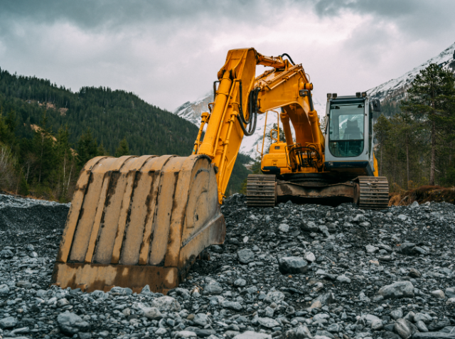 A yellow excavator on rocky terrain with mountains and a cloudy sky in the background.