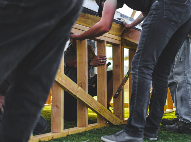 Several people collaboratively working on a wooden construction project outdoors, with a focus on hands and tools.