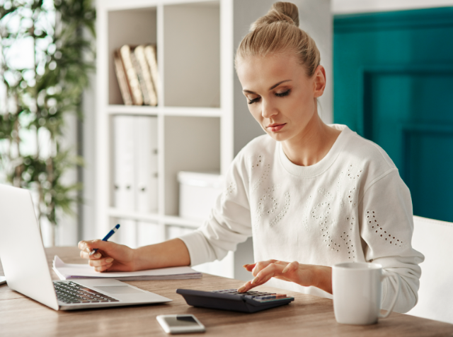 A woman calculating something using a calculator at a desk with an open laptop, smartphone, and a mug beside her in a modern room.