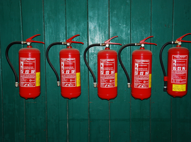 Four red fire extinguishers with hoses and safety labels mounted on a dark green wall.