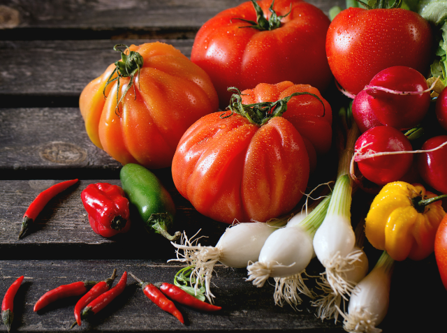 Assortment of fresh vegetables including heirloom tomatoes, cherry tomatoes, bell peppers, a jalapeño, red chili peppers, and spring onions on a wooden surface.