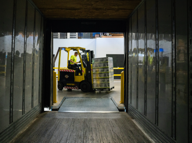 View from inside a dark cargo container towards a worker operating a yellow forklift, lifting a pallet of goods at a loading dock.