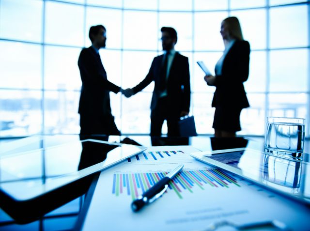 Two professionals shaking hands in a corporate setting with another observing, with business documents and a pen in the foreground.