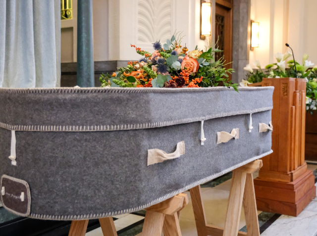 A grey felt-covered casket with white stitching and handles, adorned with a floral arrangement on top, displayed on a wooden stand inside a building with marble columns and a wooden podium.