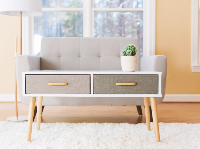 A modern living room with a beige sofa, a mid-century modern console table with a potted cactus on top, a plush white carpet, and a floor lamp, all bathed in natural light from a large window.