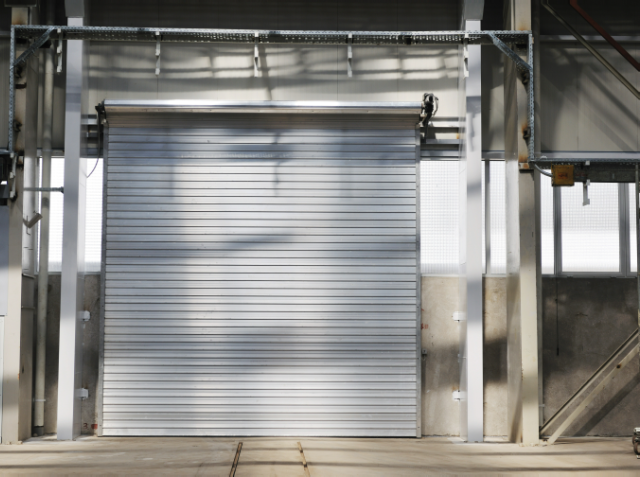 A closed roll-up metal door at the entrance of an industrial building with exposed metal beams and natural light casting shadows.