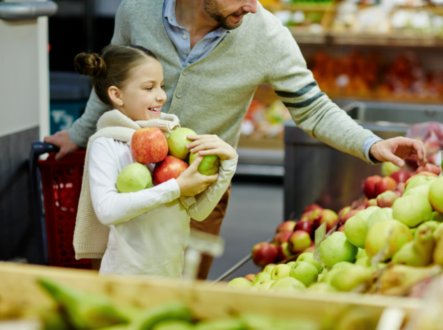 A young girl happily holding apples while an adult male, possibly her father, picks more apples from a grocery store display.