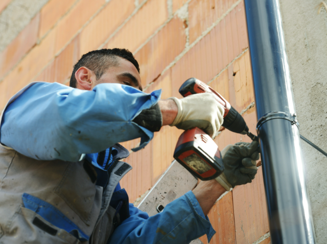 A worker in a blue uniform and gloves using a cordless drill on a pipe against a brick wall.