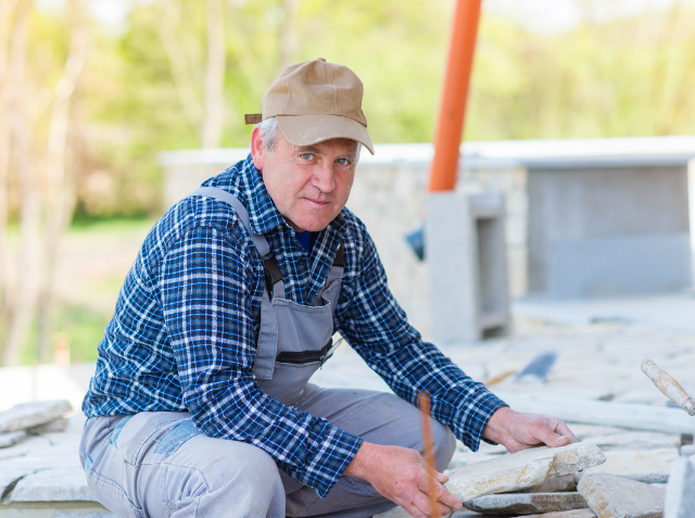 A construction worker or stonemason in a cap, plaid shirt, and overalls kneels while handling a flat stone, with an outdoor construction site in the background.