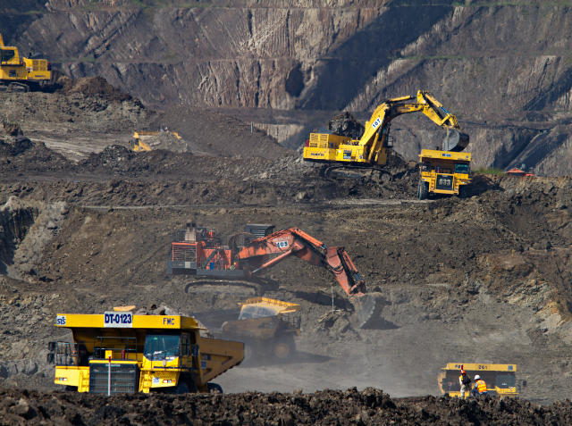 Open-pit mining operation with large yellow and red excavators and yellow dump trucks working in a dusty, terraced excavation site.