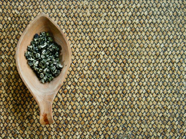 A wooden spoon filled with dried green tea leaves on a woven mat.