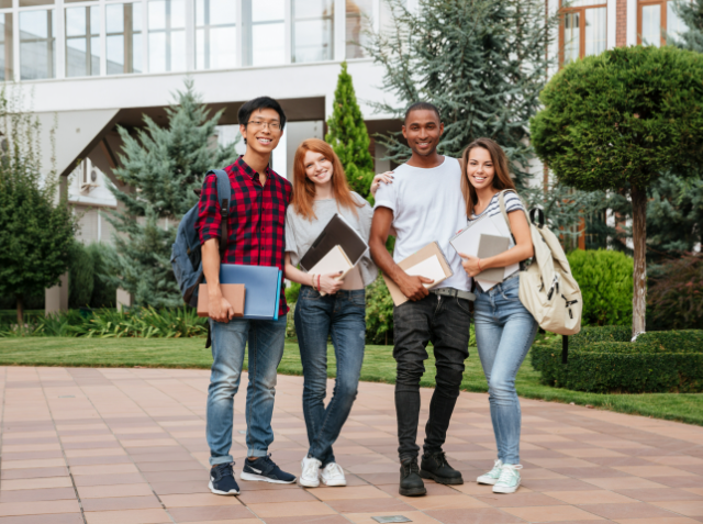 Four diverse students holding books and smiling at the camera on a university campus.