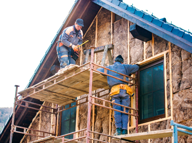 Two construction workers on scaffolding outside a stone-faced building with one measuring the exterior and the other climbing up the scaffold, both wearing hard hats and safety gear.