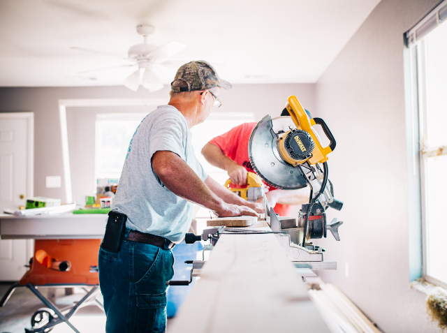 A man in casual attire using a miter saw to cut a piece of trim in an indoor setting that appears to be under renovation.